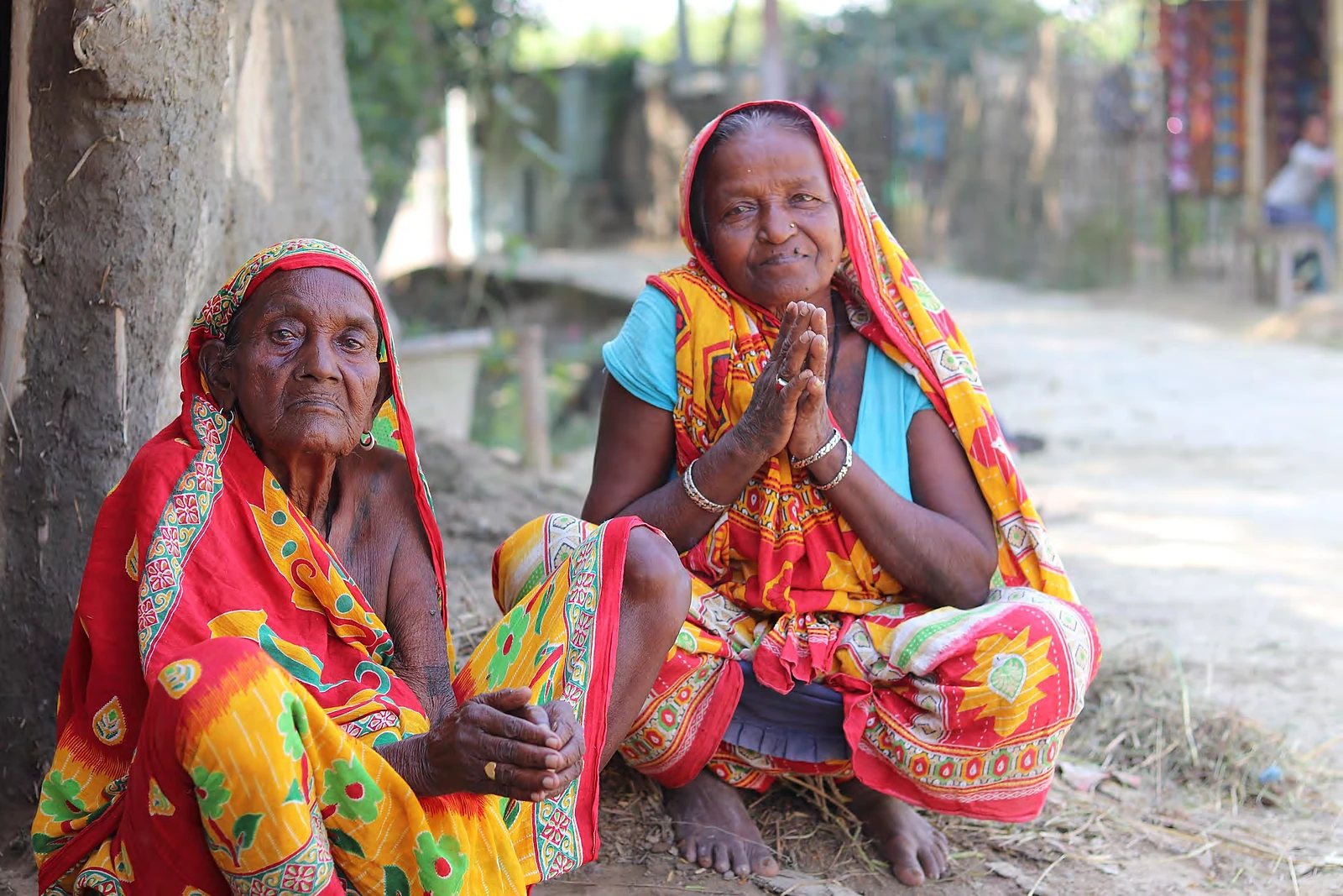Dos mujeres mayores vestidas con saris rojos y amarillos, sentadas a la sombra en un pueblo de la India